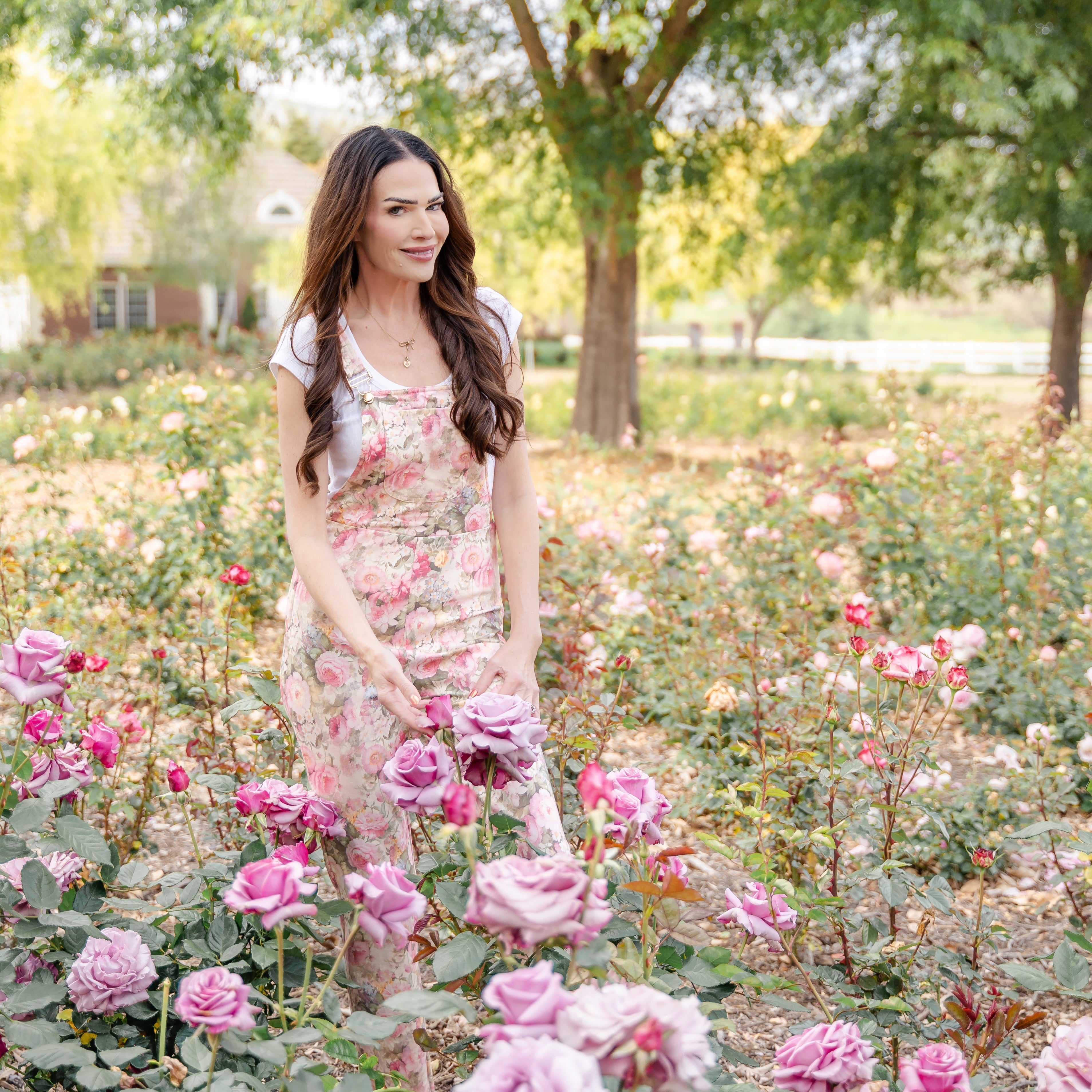 Woman in a floral dress standing among pink roses in a garden.