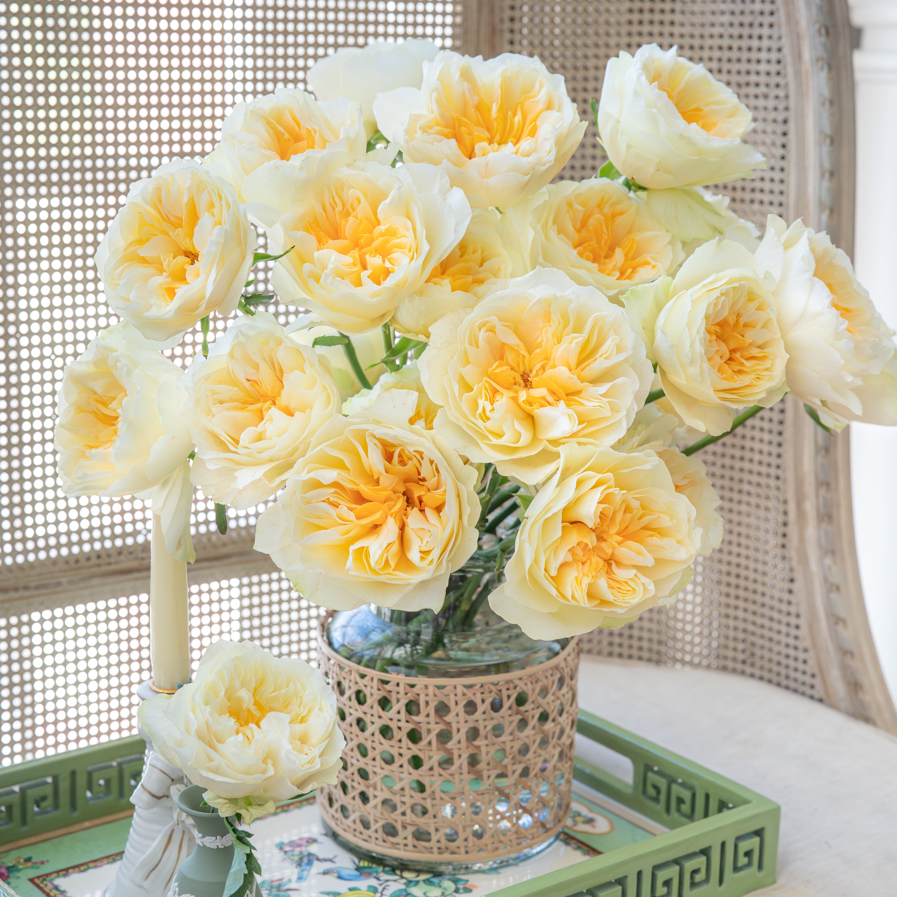 Bouquet of yellow flowers in a woven vase on a decorative tray with a wicker chair in the background.