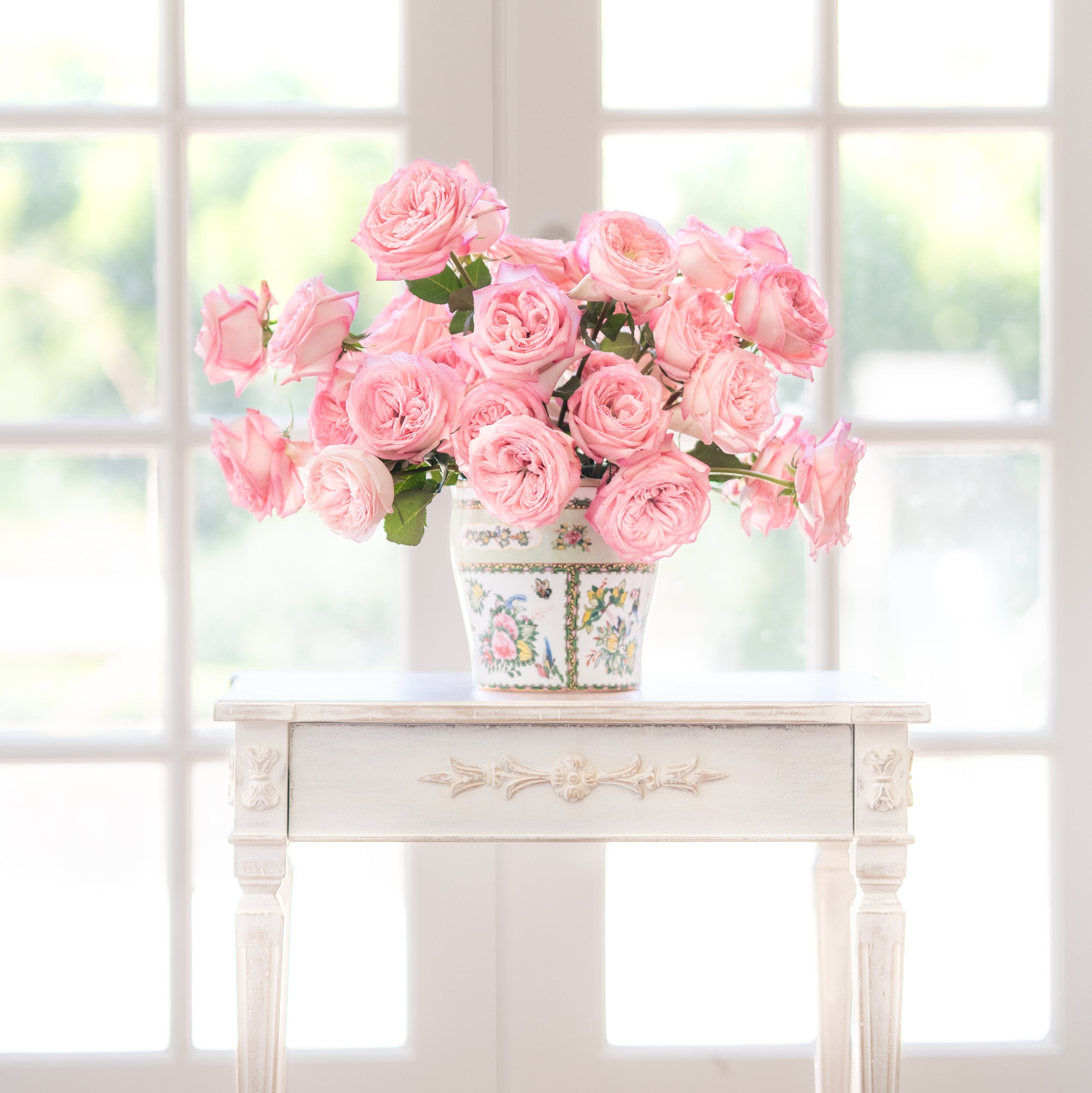 Bouquet of pink roses in a decorative vase on a white table with a window in the background