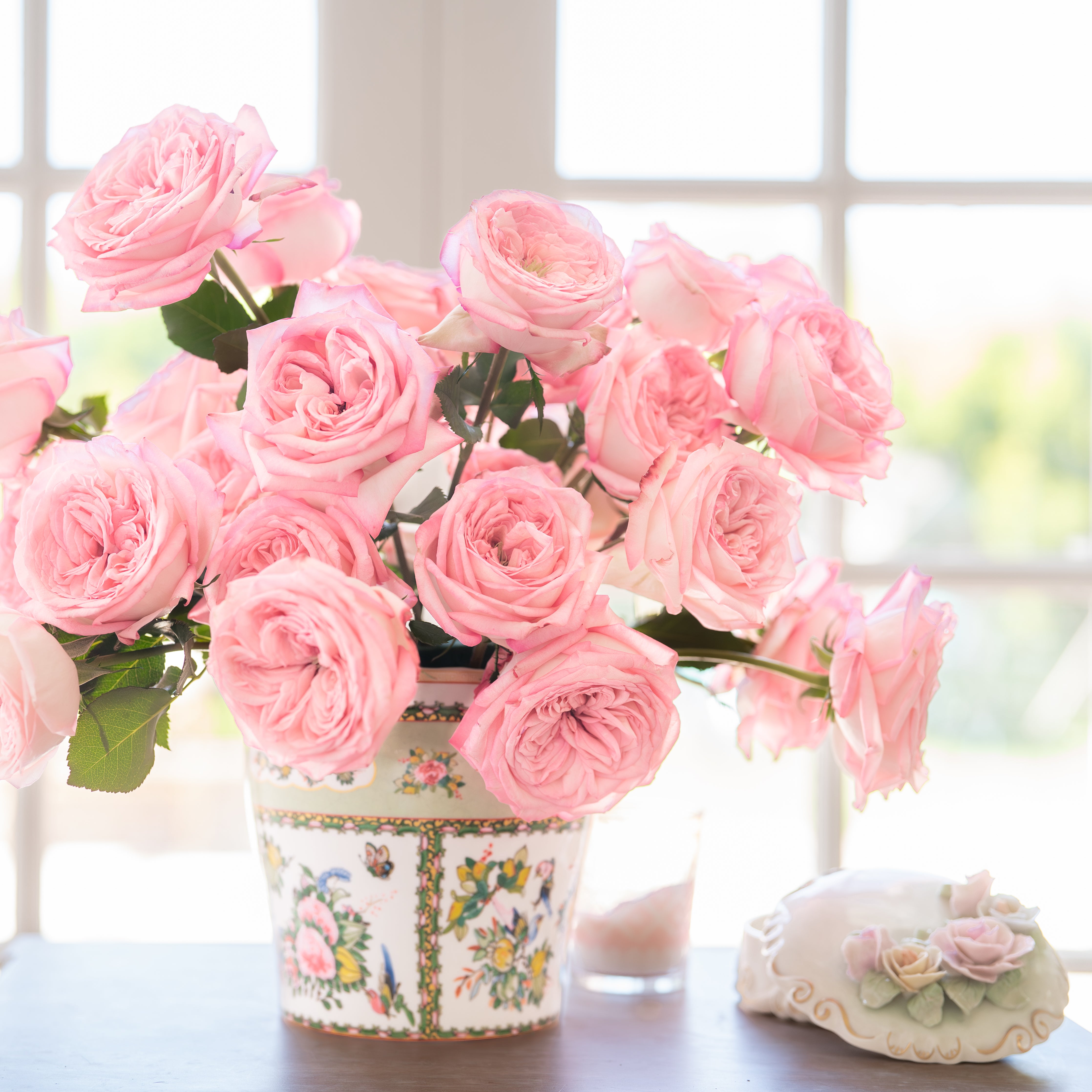Bouquet of pink roses in a decorative vase on a table with a blurred window background.